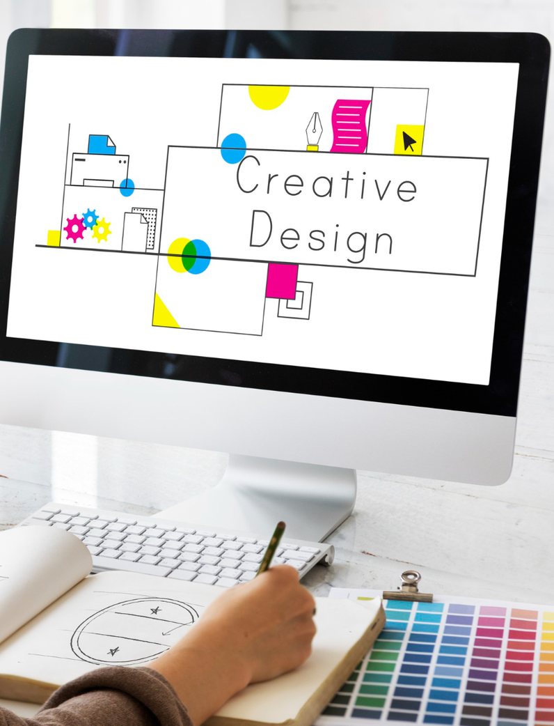 Une designer à son poste de travail : une planche de création affichée à l’écran, un carnet de croquis et un nuancier ouverts sur le bureau.