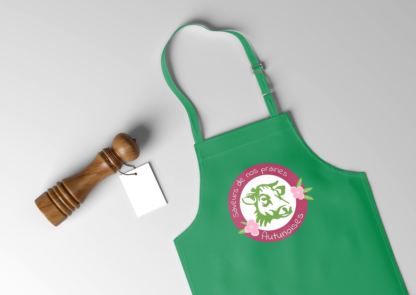 Tablier de cuisine vert imprimé du logo Saveurs de nos prairies autunoises, posé à côté d’un moulin à poivre en bois